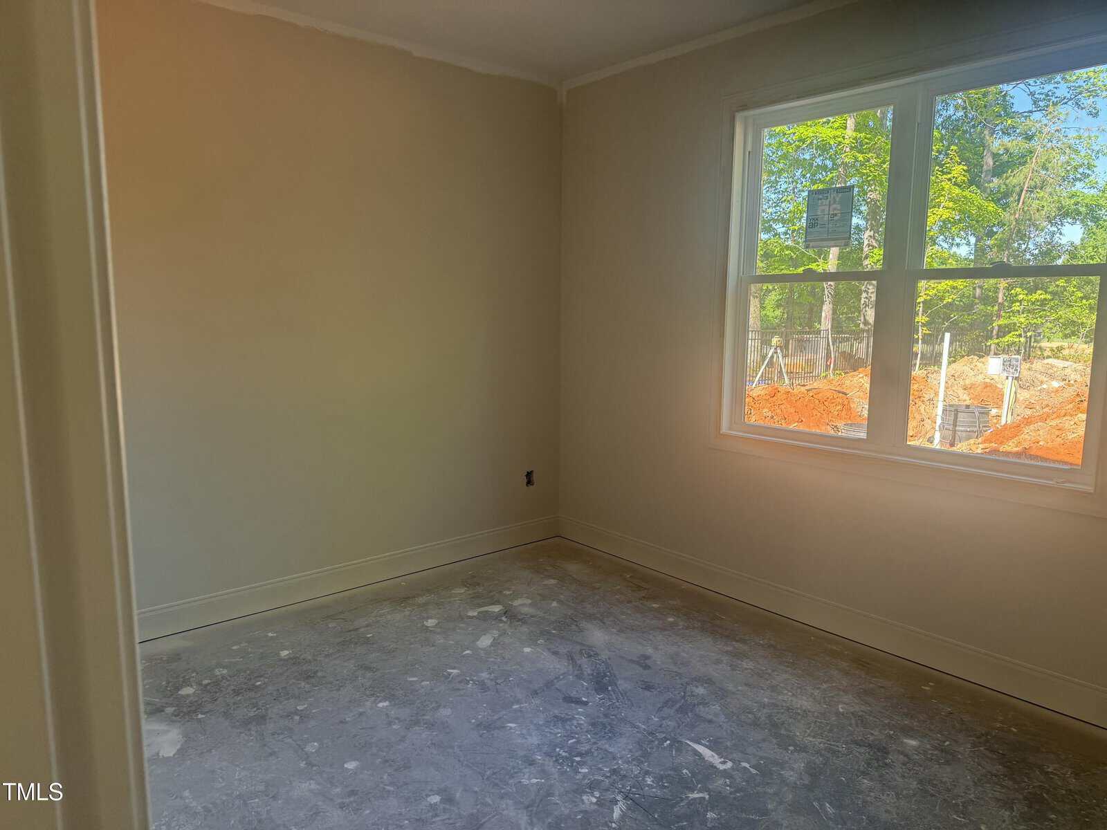 Empty, unfinished room with bare floor and beige walls. Large window lets in daylight, showing trees and construction dirt piles outside. No furniture or decorations are present.