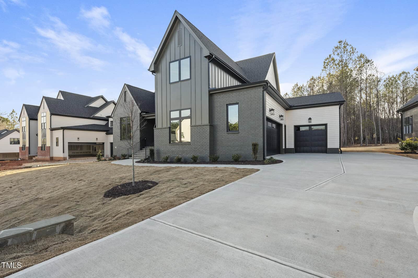 A modern two-story house with gray brick and vertical siding, black trim, a two-car garage, and a wide concrete driveway. The front yard has minimal landscaping and neighboring homes are visible on the left.
