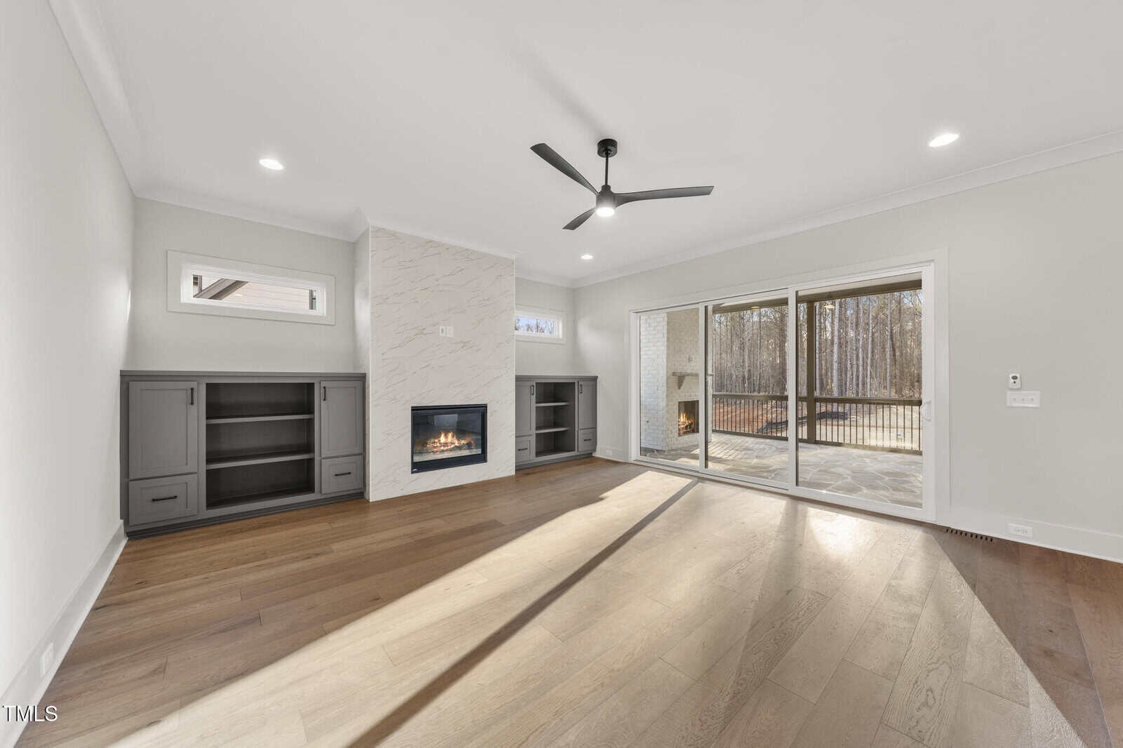 A modern living room with light walls, wood floors, a ceiling fan, built-in shelves, a fireplace with a marble surround, and large sliding glass doors leading to a patio and wooded area.