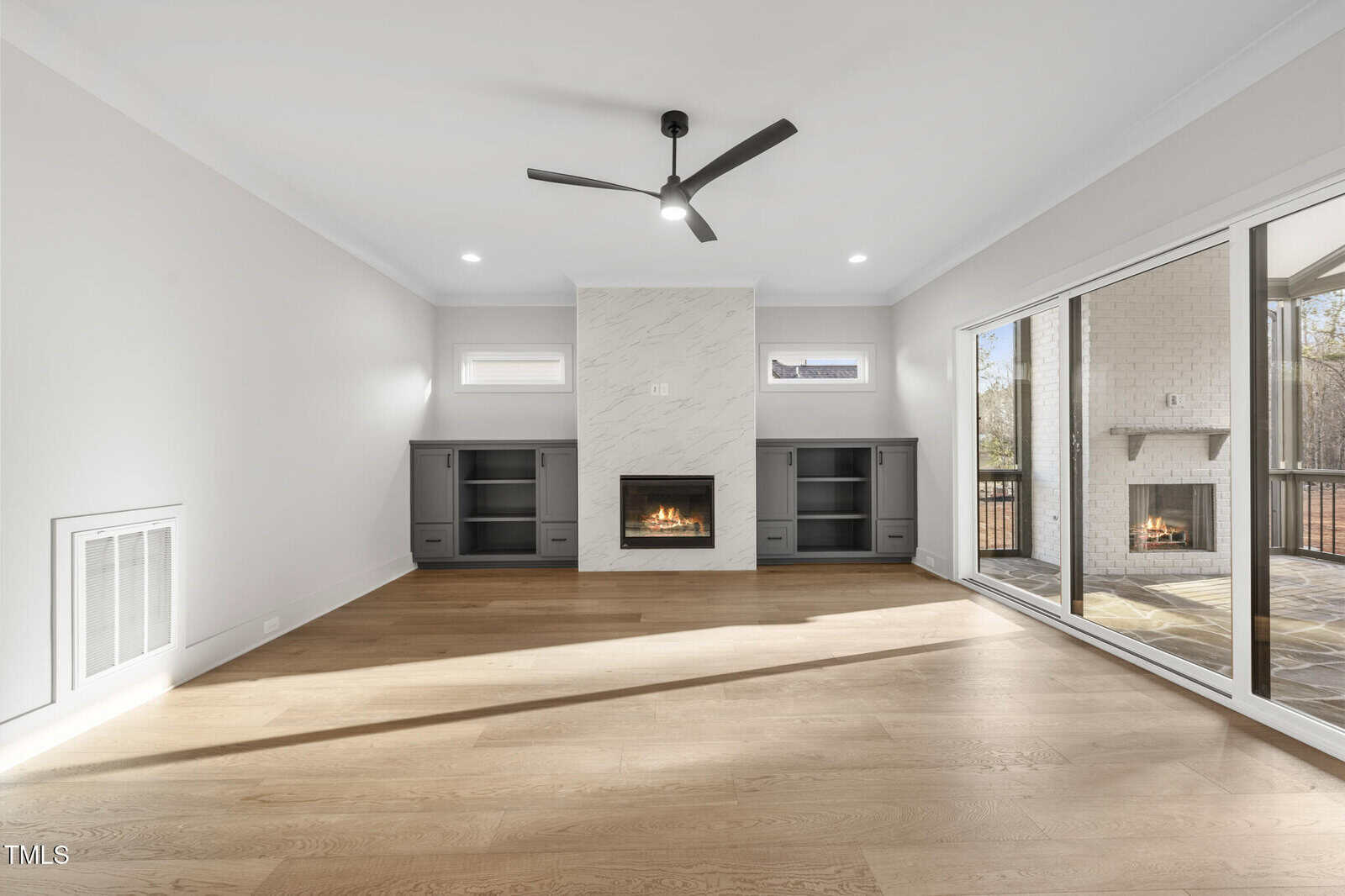 Modern living room with light wood floors, a ceiling fan, a built-in fireplace flanked by built-in shelving, two small windows, and large glass sliding doors leading to a covered patio with another fireplace.