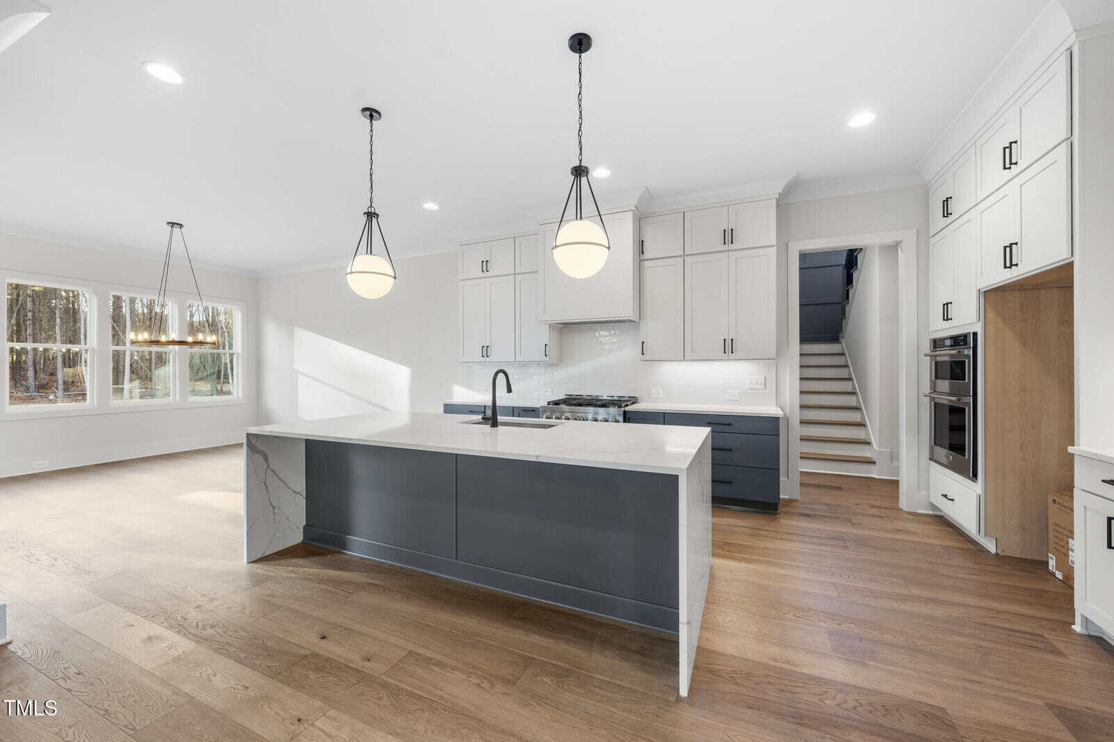 Modern kitchen with white cabinets, a gray island with a marble countertop, two pendant lights, built-in appliances, wooden floors, and a staircase leading upstairs. Large windows provide natural light.