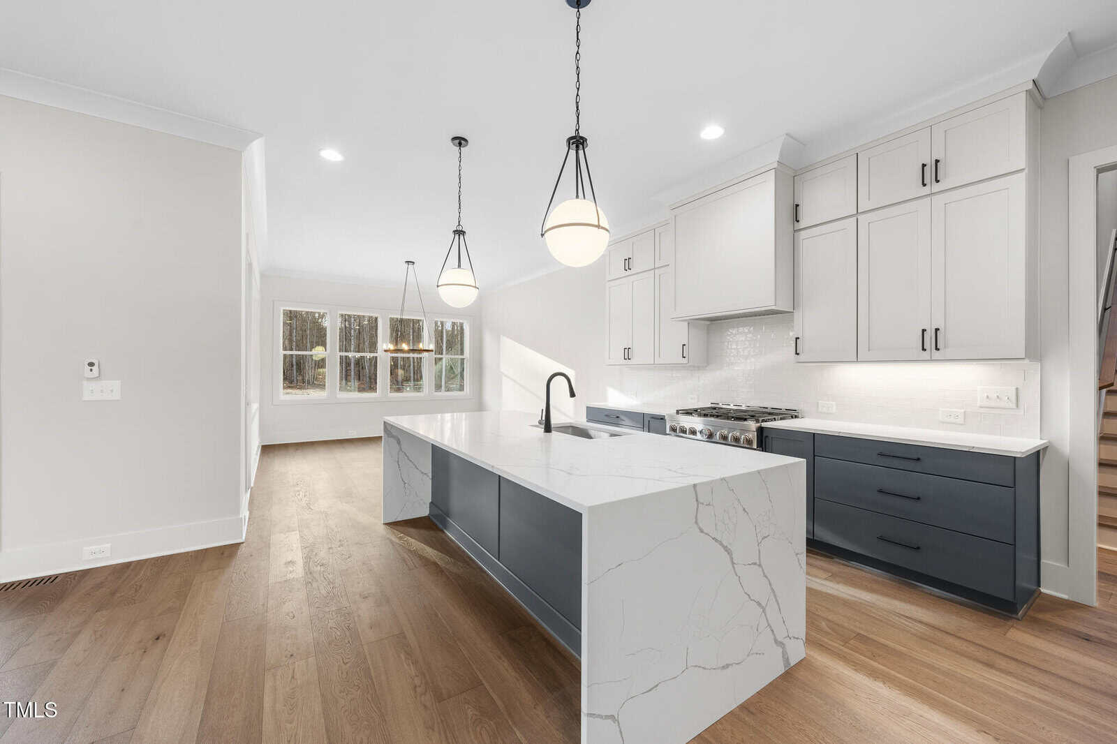 A modern kitchen with white and dark blue cabinets, a large white marble island with a sink, stainless steel appliances, pendant lights above the island, and large windows in the adjacent dining area.