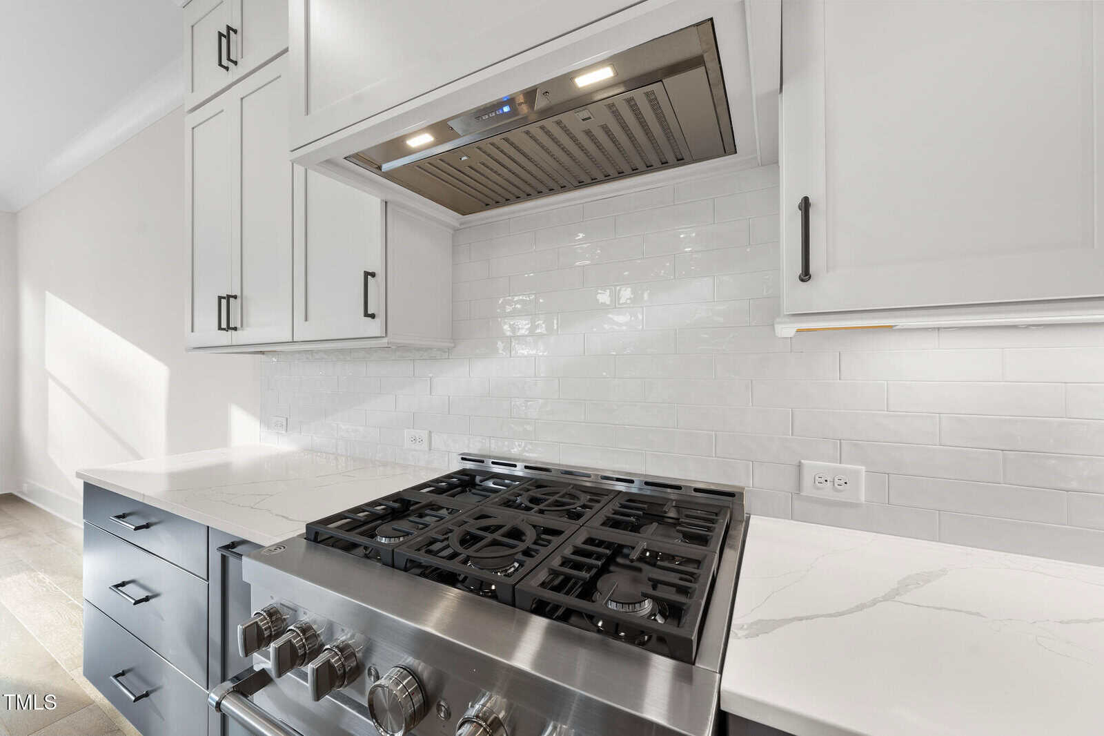 A modern kitchen features a stainless steel gas stove with multiple burners, white upper cabinets, black lower drawers, a marble-patterned countertop, and a white subway tile backsplash.