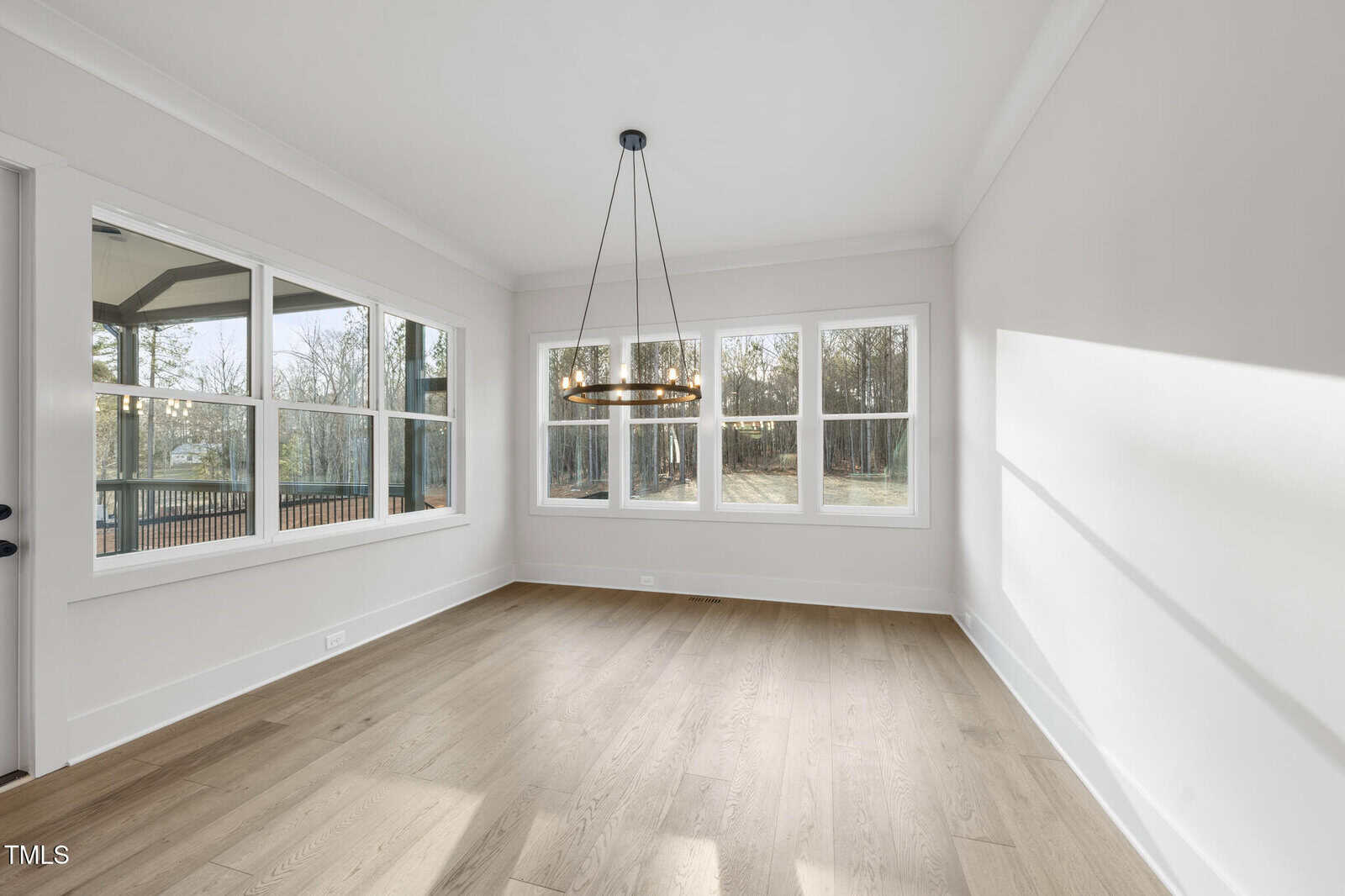 A bright, empty room with light wood floors, white walls, large windows on two sides, and a modern circular chandelier hanging from the ceiling. Sunlight streams in, highlighting the spacious interior.