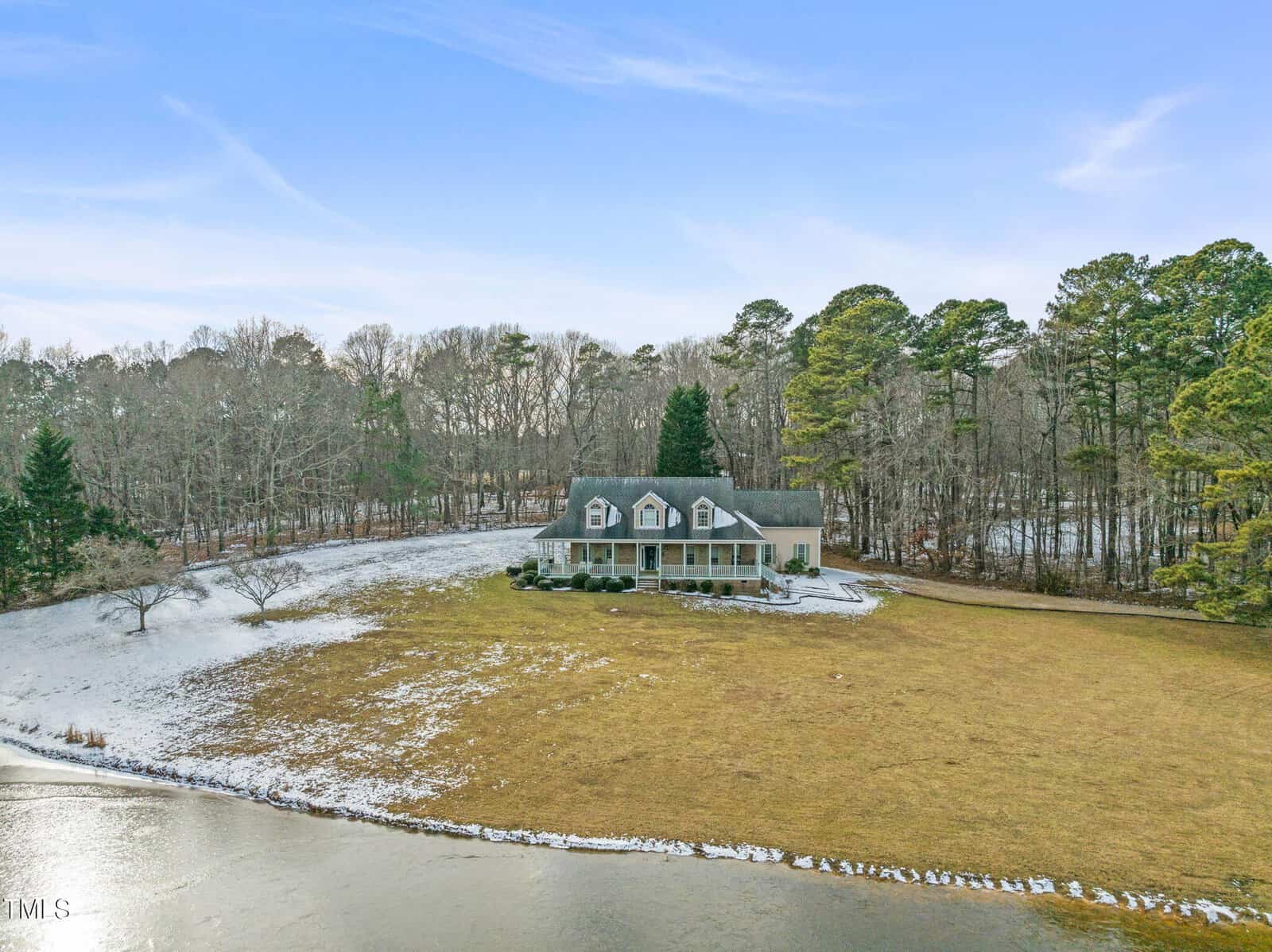 A large house at 4056 Fairfield Court sits on a grassy field surrounded by trees. A light dusting of snow covers parts of the ground and the roof. A small body of water is visible in the foreground under the clear, blue Youngsville sky.