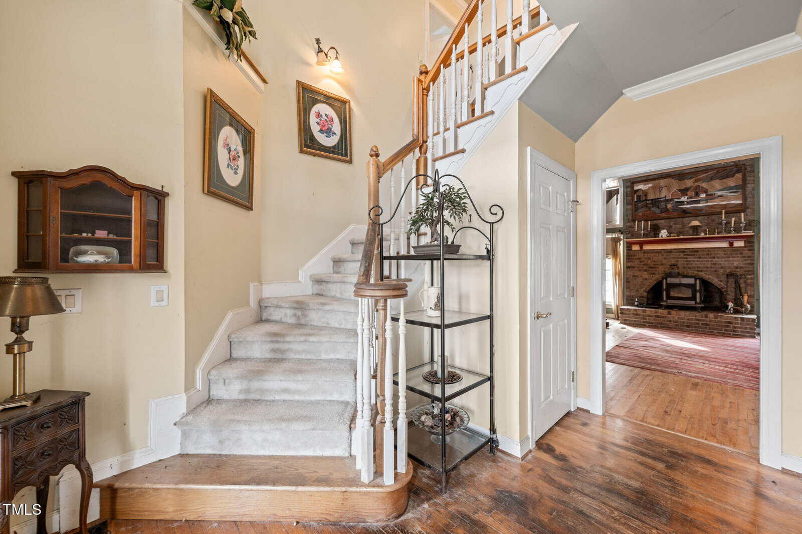 A staircase with white carpet ascends in a residential hallway. The walls are light yellow with two floral paintings. Shelves with plants are near the stairway. A doorway leads to a room with a fireplace and hardwood floor.