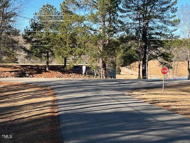 A paved road curves to meet a two-lane road with a stop sign at the intersection. Tall pine trees line both sides of the road, and power lines are visible overhead under a clear blue sky.