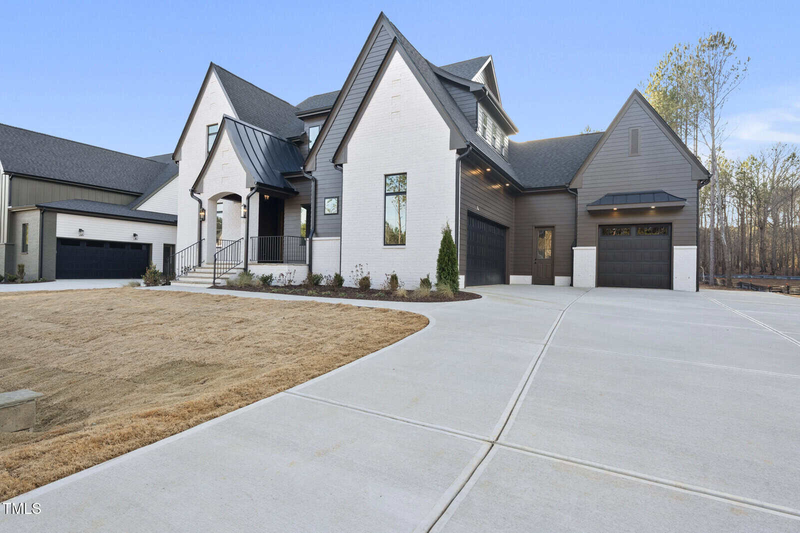 Modern two-story house with a mix of white brick and dark siding, black garage doors, large windows, and a wide concrete driveway. The front yard has minimal landscaping and the sky is clear.