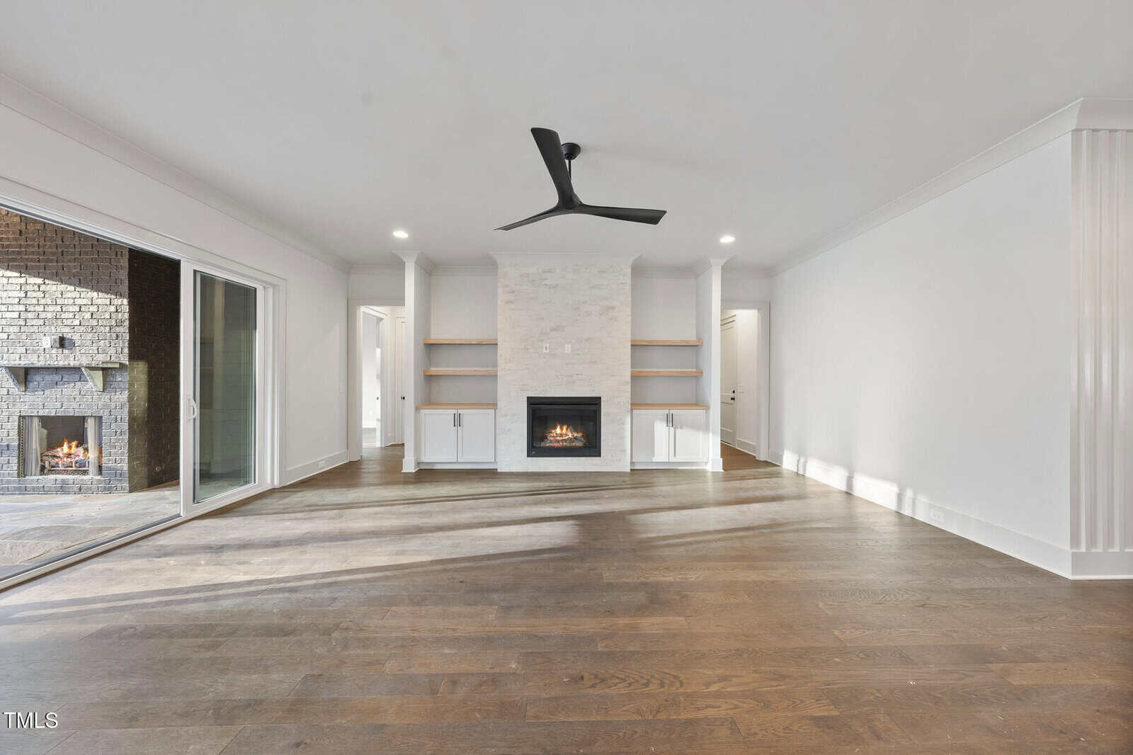 Spacious living room with wood flooring, white walls, a black ceiling fan, fireplace with white mantel, built-in shelves, and sliding glass doors leading to a patio with a second fireplace.
