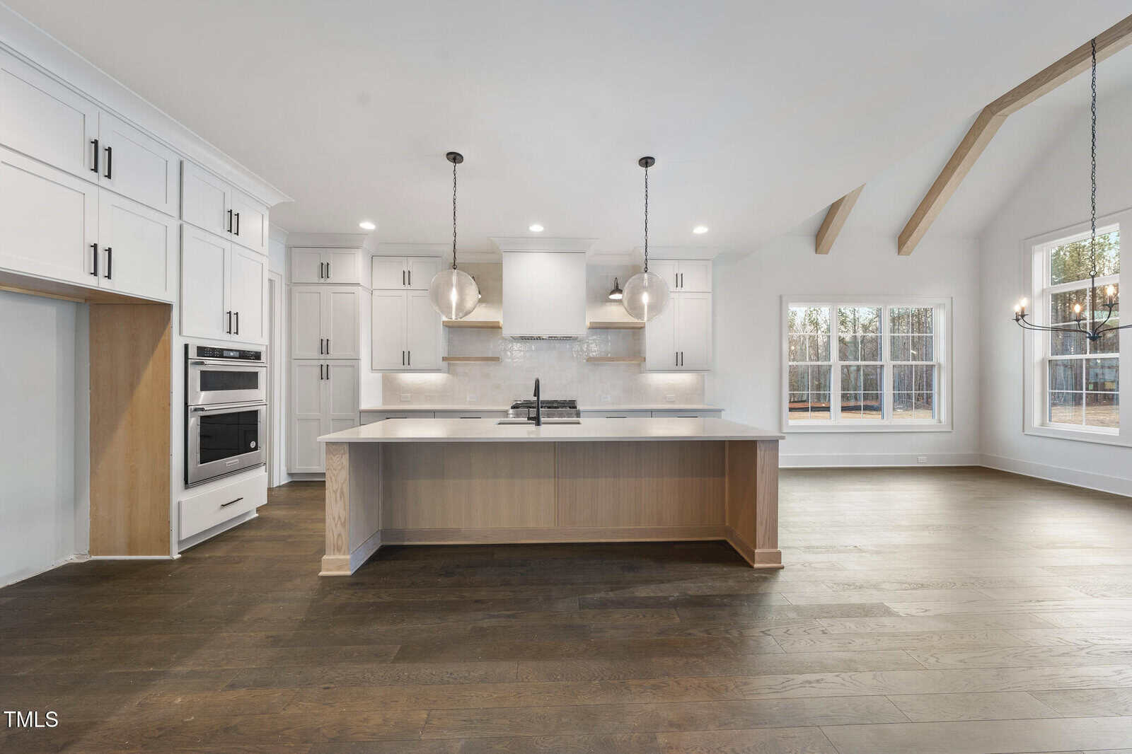Modern kitchen with white cabinets, a large central island with wood accents, stainless steel appliances, pendant lights, and hardwood floors. Large windows let in natural light; the space is empty of furniture.