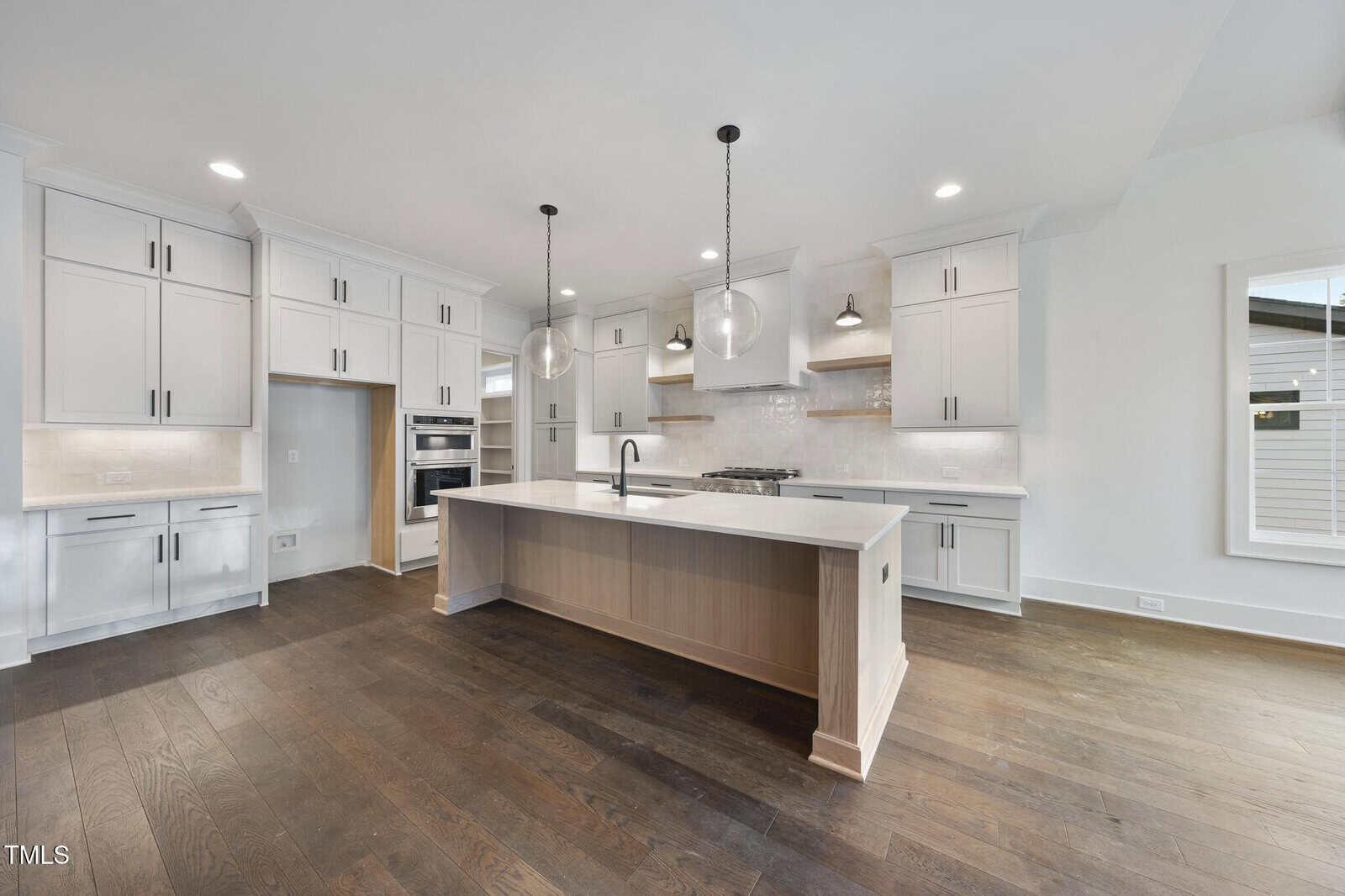 Spacious modern kitchen with white cabinetry, stainless steel appliances, a large center island with sink, pendant lights, and dark wood flooring. Room features white walls and ample natural light from windows.
