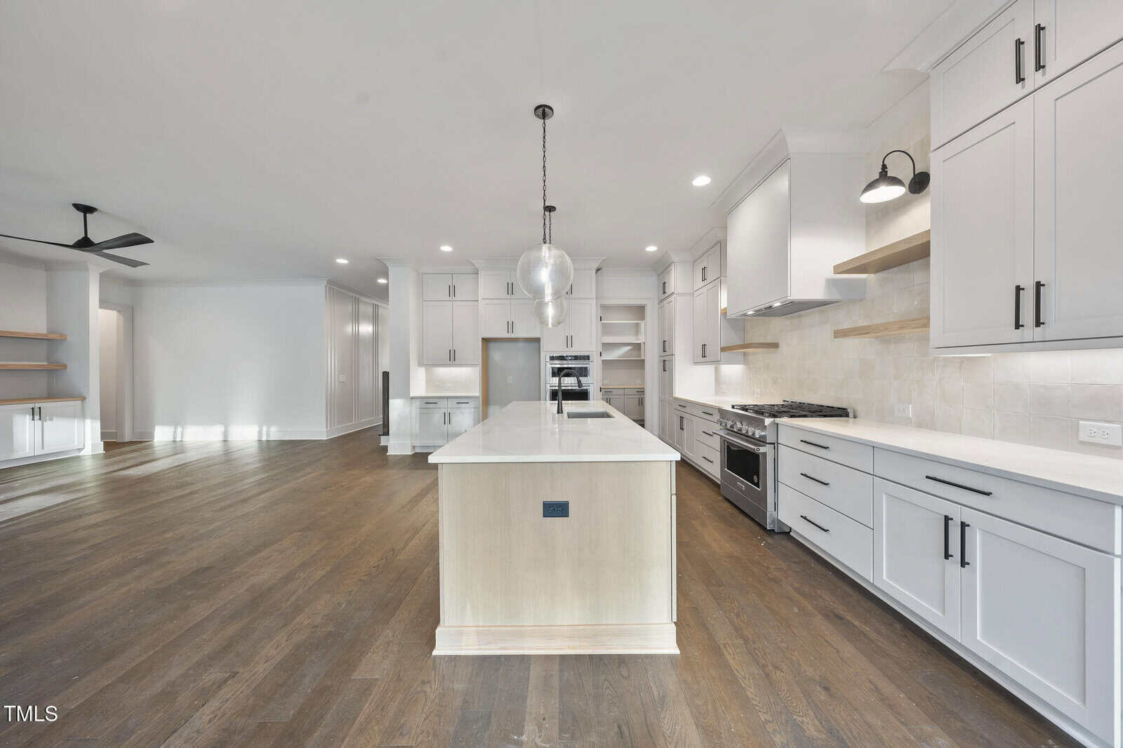 Spacious modern kitchen with white cabinets, stainless steel appliances, a large center island with a pendant light, dark wood floors, open shelving, and a ceiling fan in the adjacent living area.