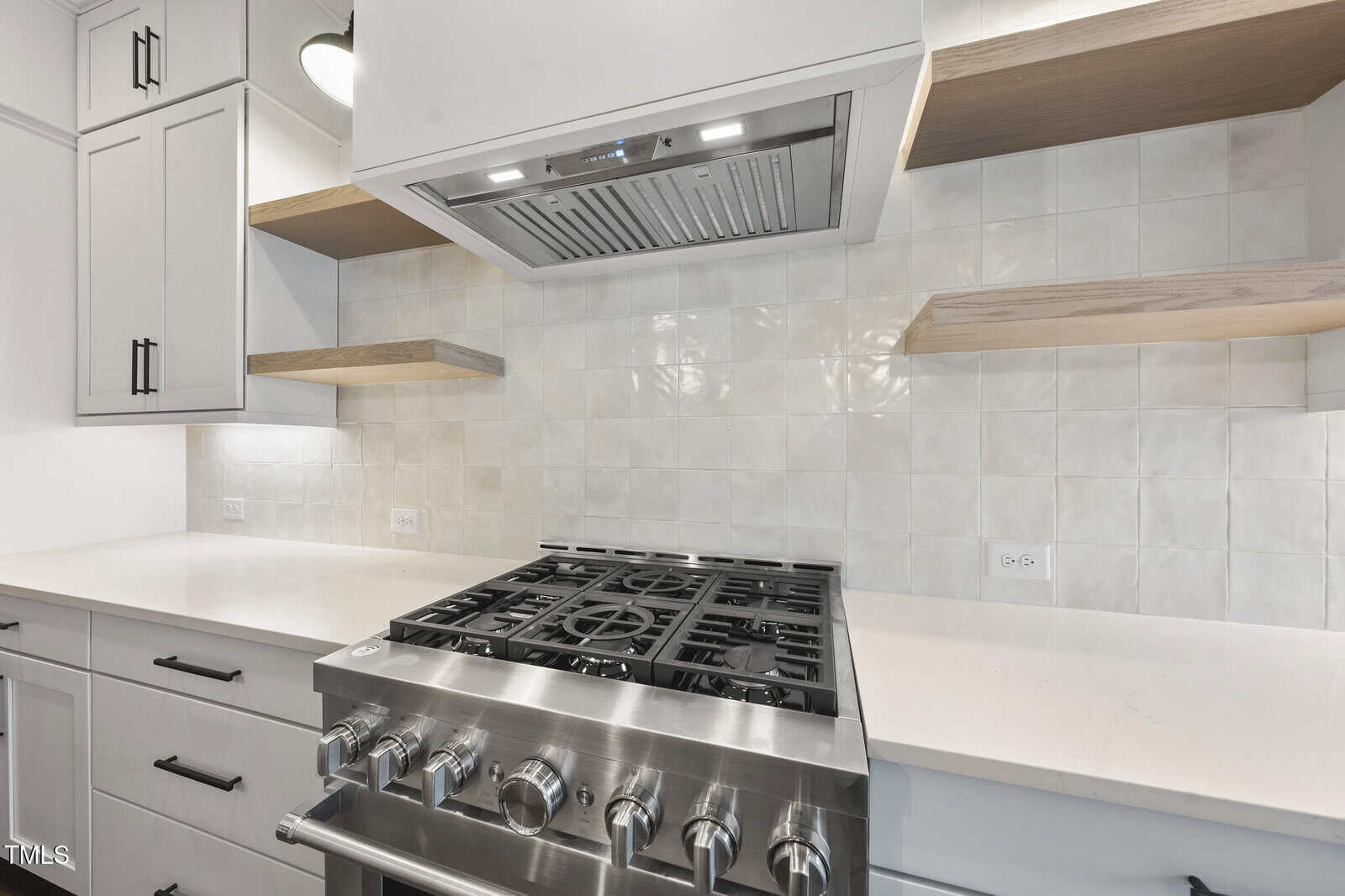 A modern kitchen features a stainless steel gas range with five burners, white tile backsplash, white countertops, light wood floating shelves, and white cabinetry with black handles.
