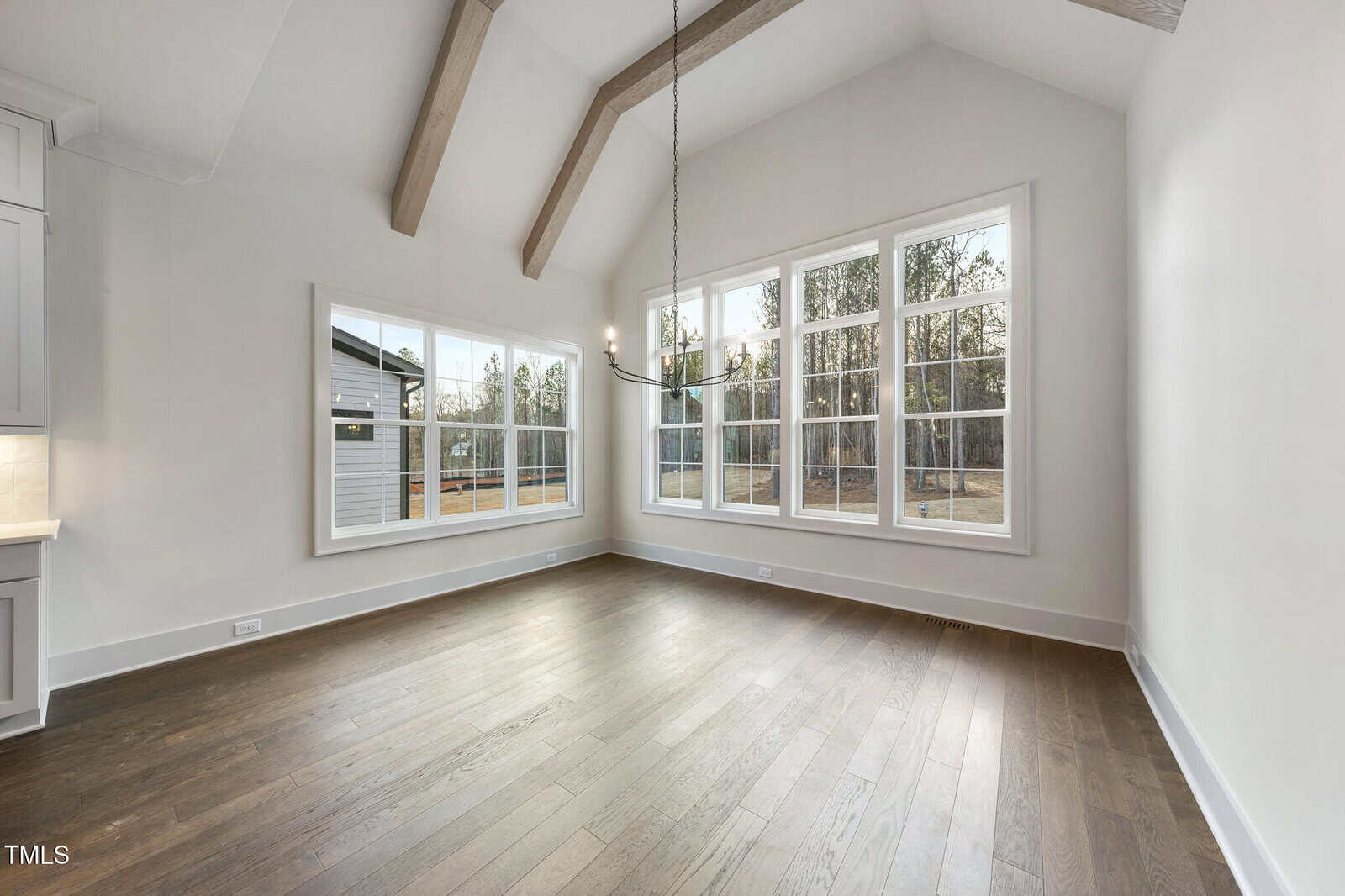 Bright, empty room with large windows, wood floors, vaulted ceiling with exposed beams, and a modern chandelier. Sunlight comes in through the windows, and trees are visible outside. Part of a kitchen is visible on the left.