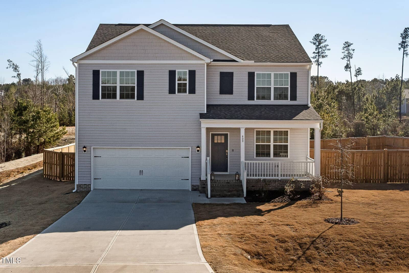 Two-story light gray house with black shutters, attached double garage, front porch with white railings, concrete driveway, and a large fenced backyard. The lawn is dry with a few small trees visible.