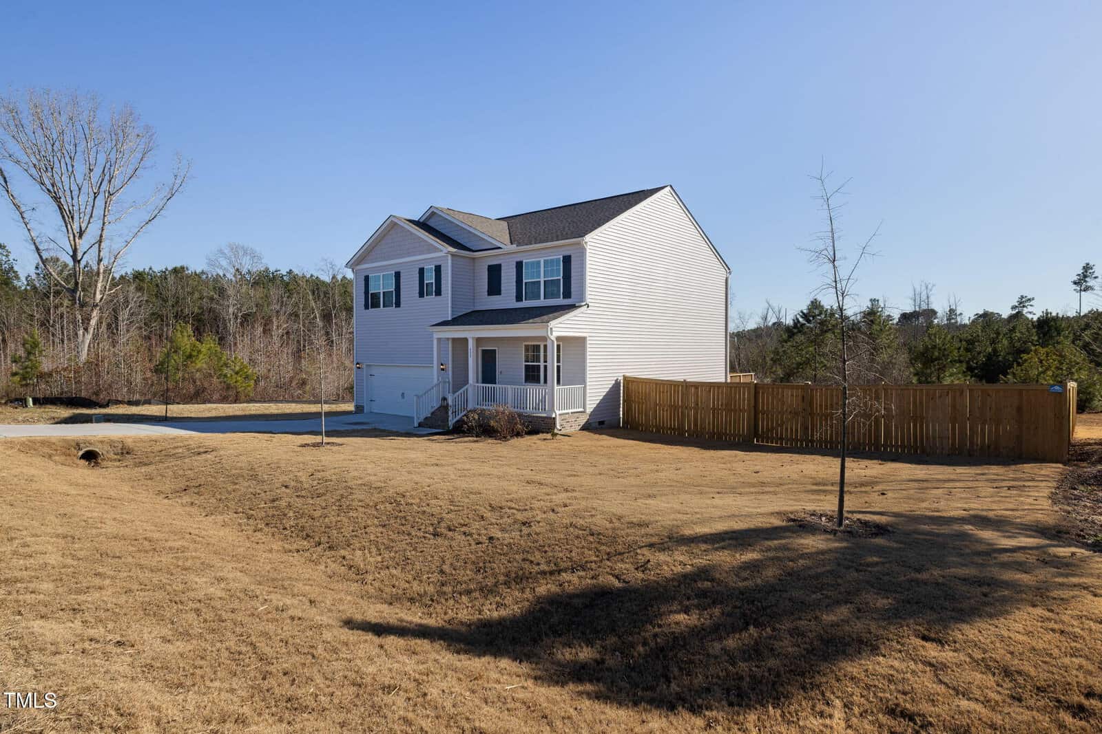 A two-story white house with a covered front porch and attached garage sits on a large lawn with sparse grass. A wooden privacy fence borders part of the backyard, and trees are visible in the background under a clear sky.
