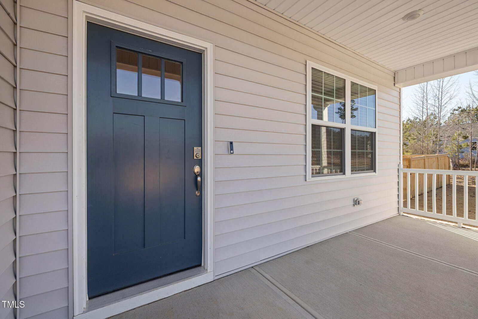 A covered porch with a blue front door, beige siding, a large window, and a white railing. The porch floor is concrete and a residential yard with trees is visible in the background.
