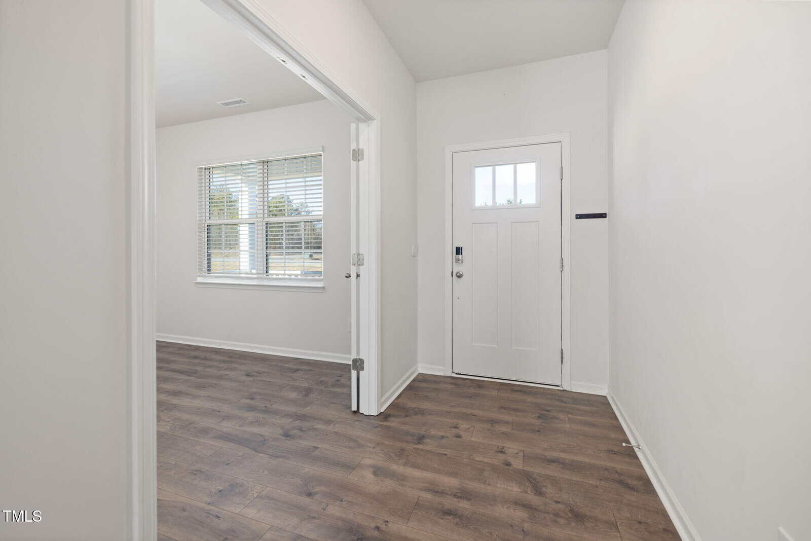 A white front door with a window is shown in a bright entryway with light-colored walls and wood-style flooring. An adjacent room with double doors and a window is visible on the left.