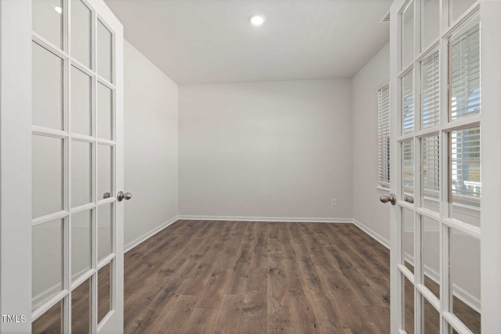 A small empty room with white walls, wood flooring, and double glass doors. One wall has two windows with blinds. A single recessed ceiling light is on. The room appears clean and well-lit.
