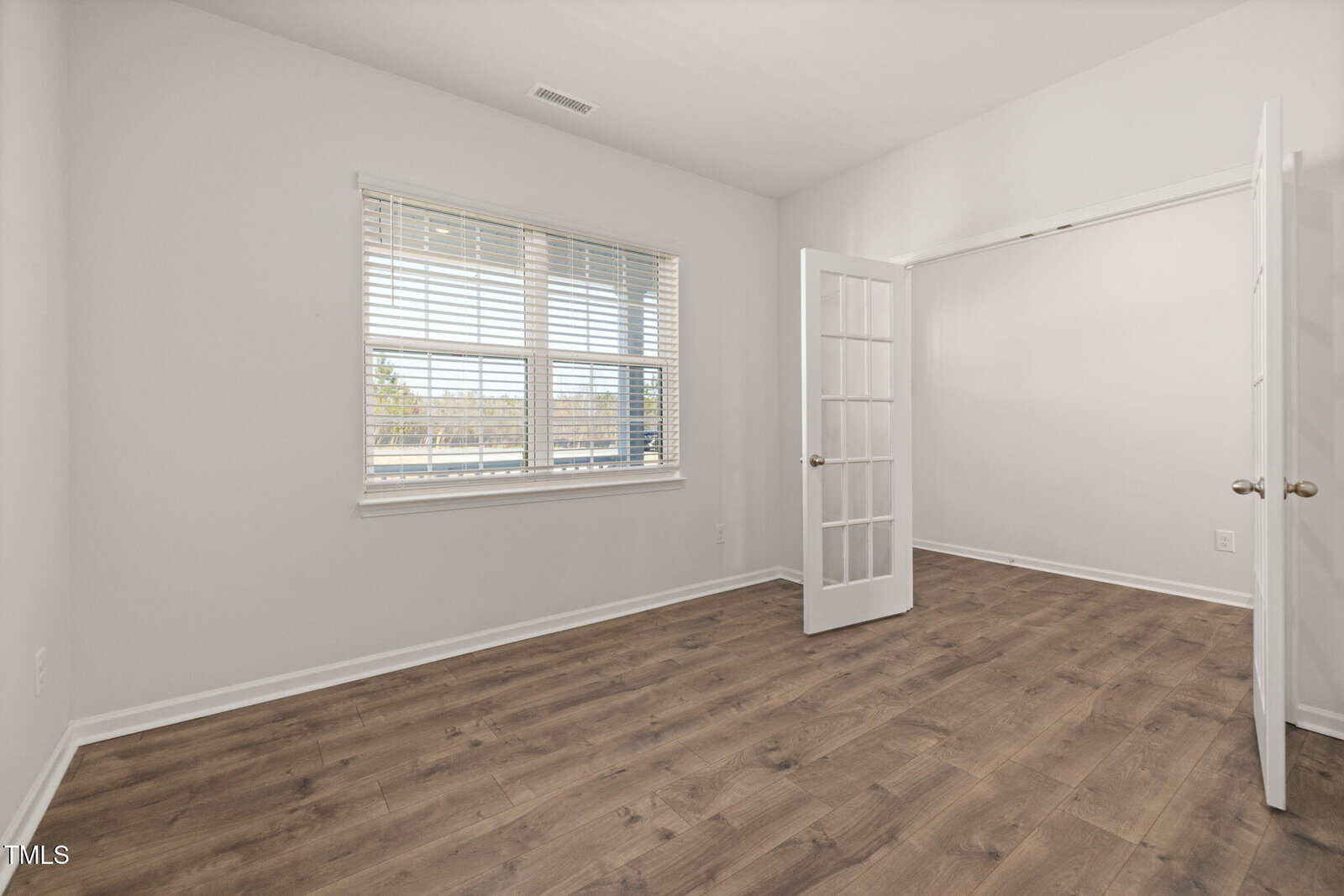 Empty room with light-colored walls, a large window with blinds, wood-style flooring, and double glass French doors open to an adjacent space. Natural light fills the room.