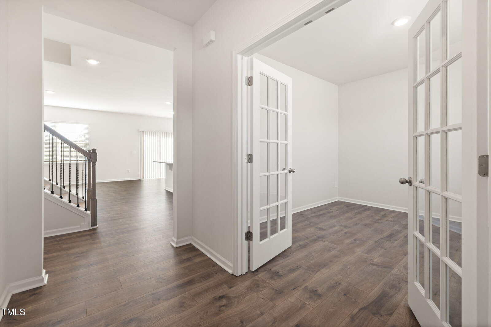 A bright, empty room with double glass French doors opens to a hallway with dark wood flooring. Stairs with a black railing are visible on the left, and sunlight filters through blinds in the background.