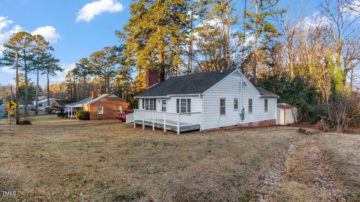 A small white house with a front porch sits on a grassy yard at 902 Roanoke Avenue in Henderson. Tall trees surround the area, and a brick house is visible in the background. The sky is clear with a few clouds, adding to the serene charm of the neighborhood.