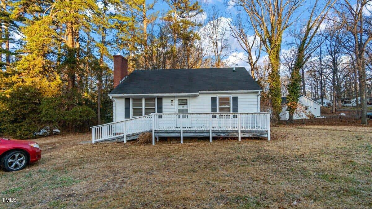 A quaint, single-story white house with a dark roof and a chimney sits on a grassy yard. It features a white ramp leading to the front door. The background has tall trees and a partly cloudy sky. A red car is parked on the left side of the house.