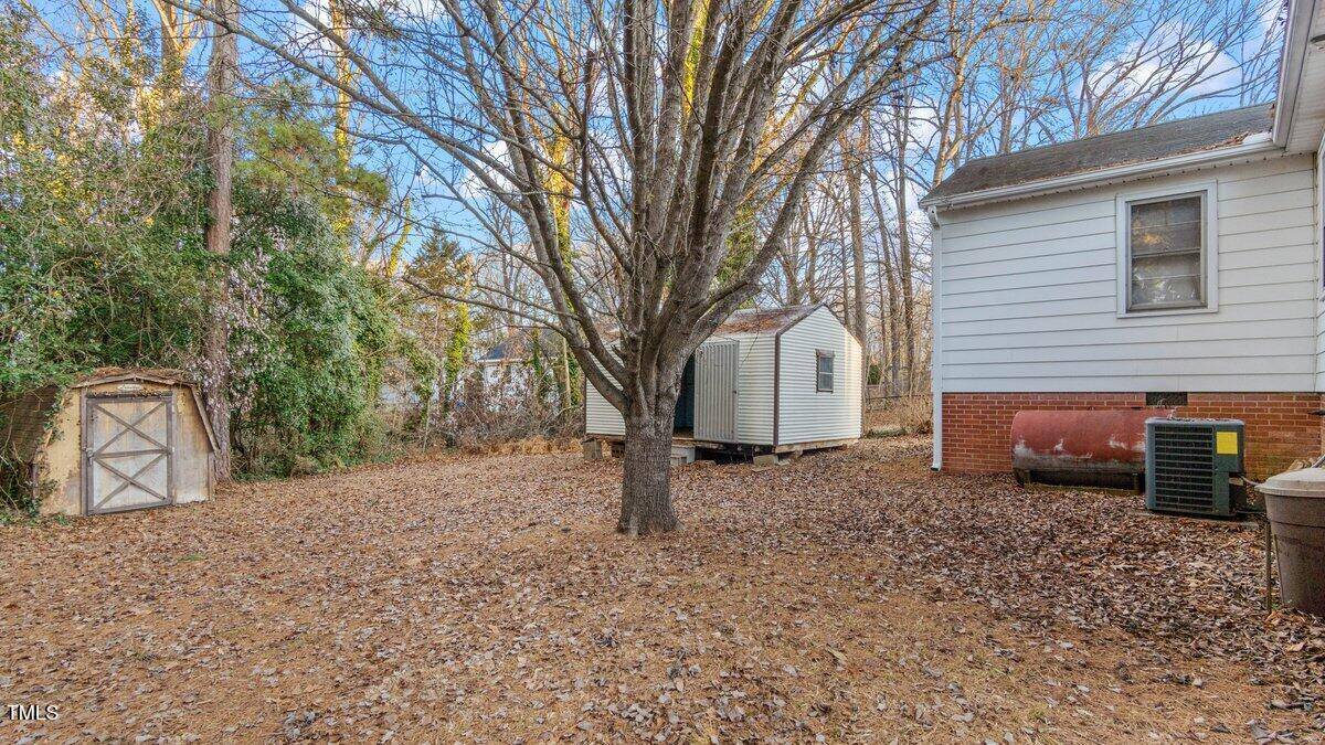 A backyard with a central tree surrounded by fallen leaves. Two small sheds are present to the left and center. A portion of a white house with a red brick foundation and an HVAC unit is on the right. Bare branches are visible against the sky.