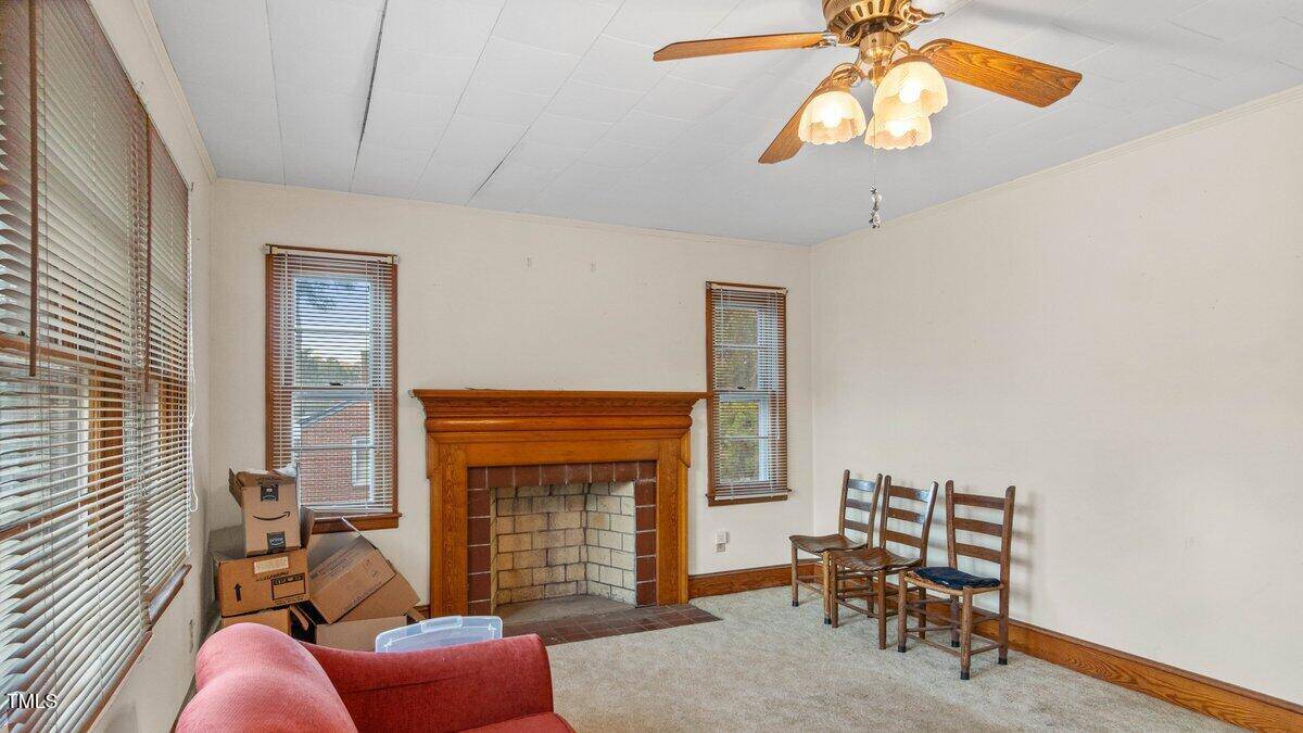 A small, partially furnished room with a ceiling fan. There's a red sofa, three wooden chairs, and a brick fireplace. A few cardboard boxes and a plastic bin are on the floor. Windows with blinds allow natural light in.