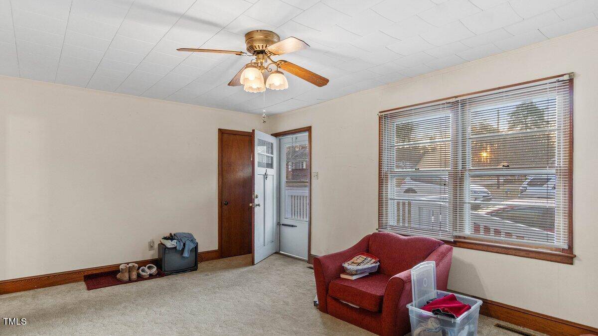 Living room with beige carpet, cream walls, and a ceiling fan with lights. A red armchair sits by a window. An open door leads outside. Shoes and bags are near the doorway. A plastic container and books are on the floor.