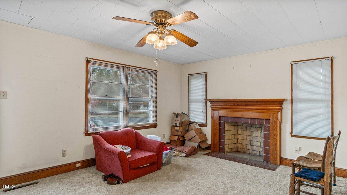 Living room with a red armchair, a brick fireplace with a wooden mantel, brown cardboard boxes, and two wooden chairs. A ceiling fan with lights is centrally mounted, and there are three windows with blinds. The walls are beige and floor is carpeted.