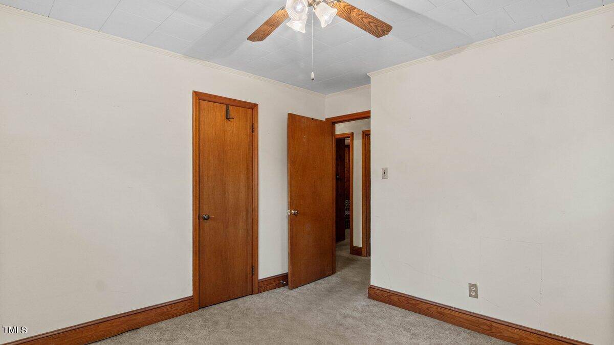 An empty room with beige walls and a beige carpeted floor. There is a wooden door slightly ajar and a closed wooden closet door. A ceiling fan with lights is attached to the white ceiling.