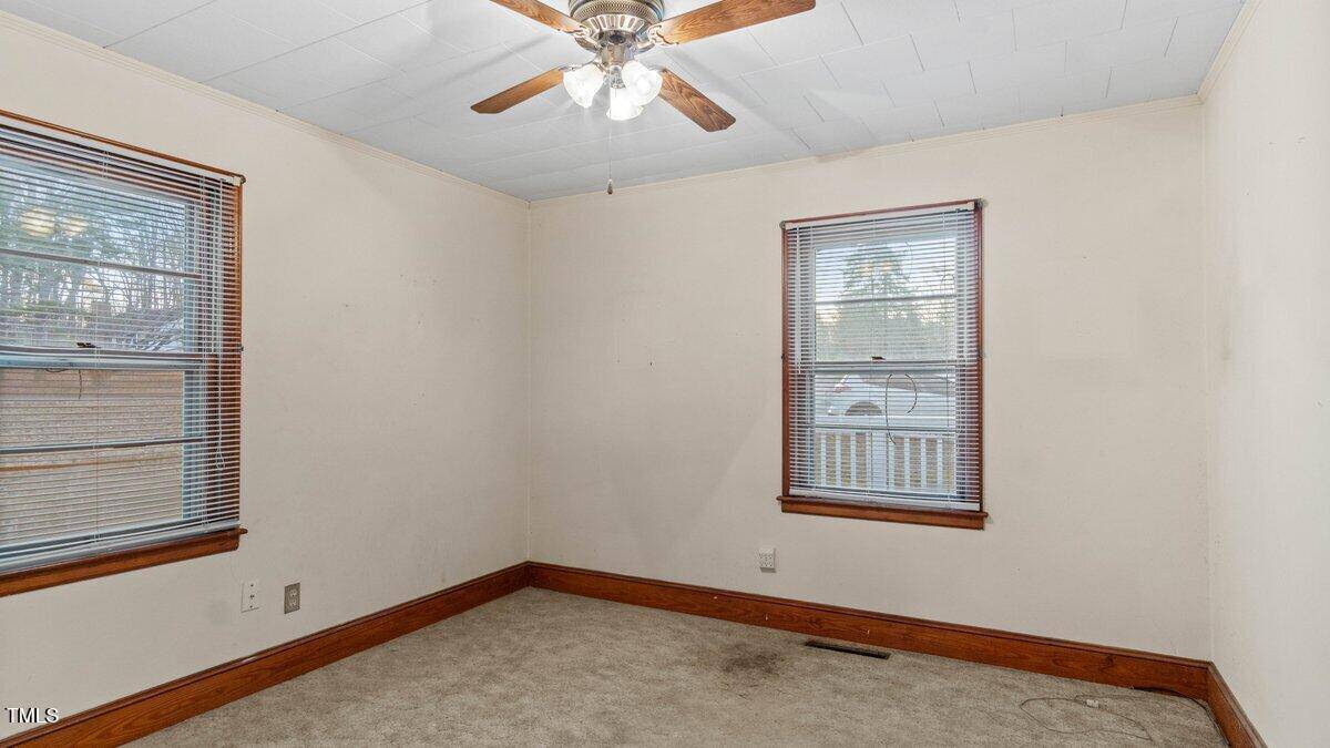 A small, empty room with beige walls and beige carpet. Two windows with wooden trims offer outside views. A ceiling fan with lights hangs from the white ceiling. The room appears minimally furnished and well-lit.