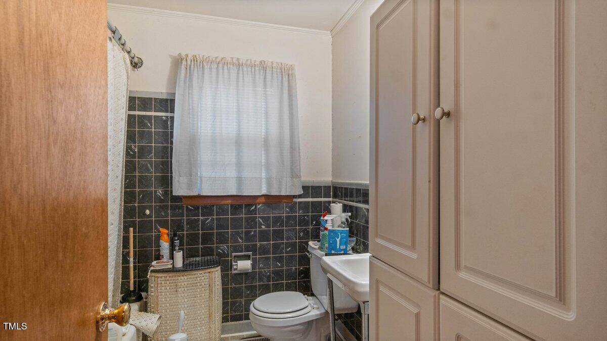 A small bathroom featuring a tiled wall with a window covered by a sheer curtain. There is a toilet, a pedestal sink with toiletries, a wicker basket, and a cabinet. A shower curtain is partially visible on the left side.