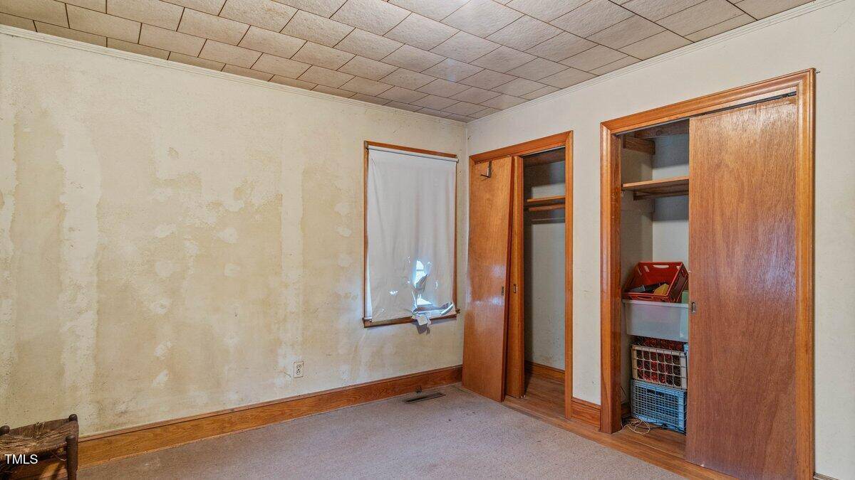 A small, empty room with beige walls featuring peeled paint. There is a single window with a wrinkled white curtain. Two open wooden closets reveal shelves and hangers. The ceiling is tiled, and the floor is carpeted.