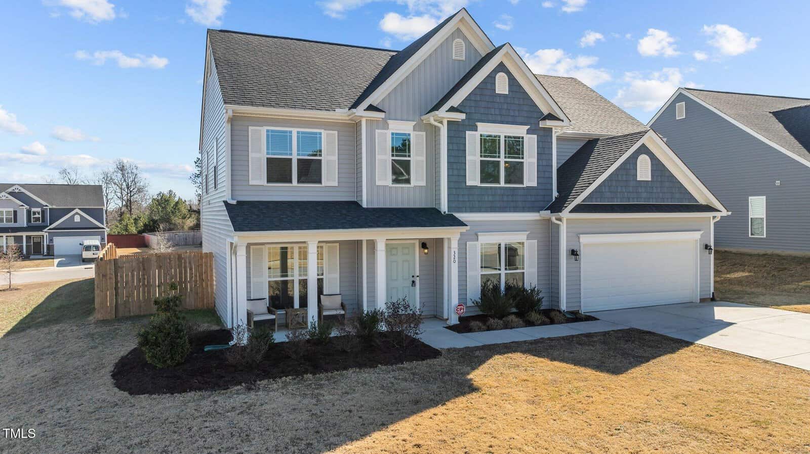 A two-story suburban house with gray siding, white trim, and a two-car garage sits on a corner lot. The front porch has columns and landscaping with shrubs. A wooden fence separates the backyard. The sky is clear with scattered clouds.