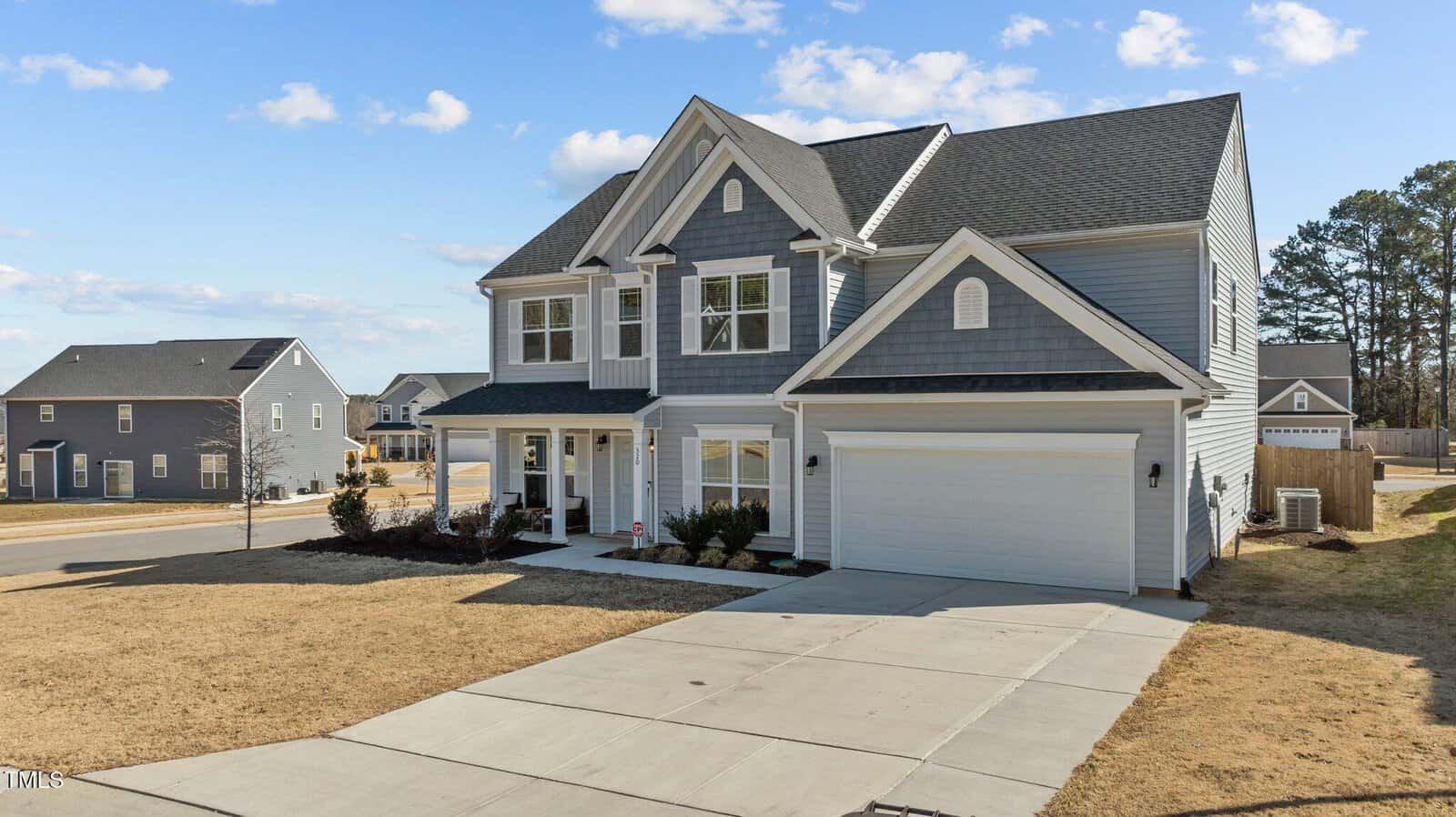 A two-story gray house with white trim, a covered front porch, double garage, and a large concrete driveway sits on a corner lot with a dry, yellowed lawn under a blue sky with scattered clouds.
