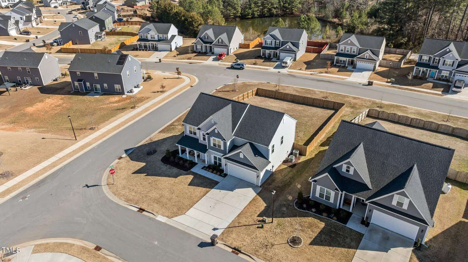 Aerial view of a suburban neighborhood featuring two-story and single-story houses, wide streets, driveways, brown lawns, and fenced backyards, with trees and a pond in the background.