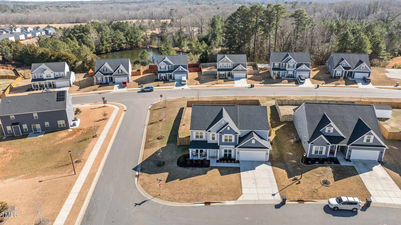 A suburban neighborhood features two-story houses with driveways, garages, and neatly maintained yards lining a curved street. There are cars parked in some driveways, and the area is surrounded by trees and a small pond.