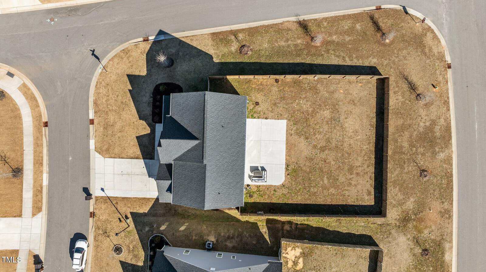 Aerial view of a single house with a dark roof, driveway, fenced backyard, and some trees. The property is bordered by roads on two sides in a suburban neighborhood. A white car is parked on the street.