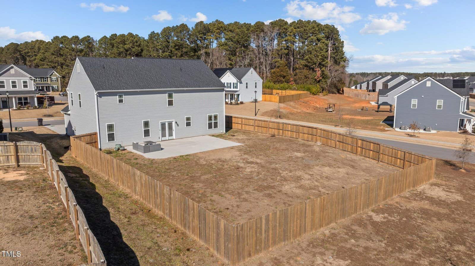 A two-story gray house with a fenced backyard featuring a concrete patio, situated in a suburban neighborhood with similar houses and some trees in the background.