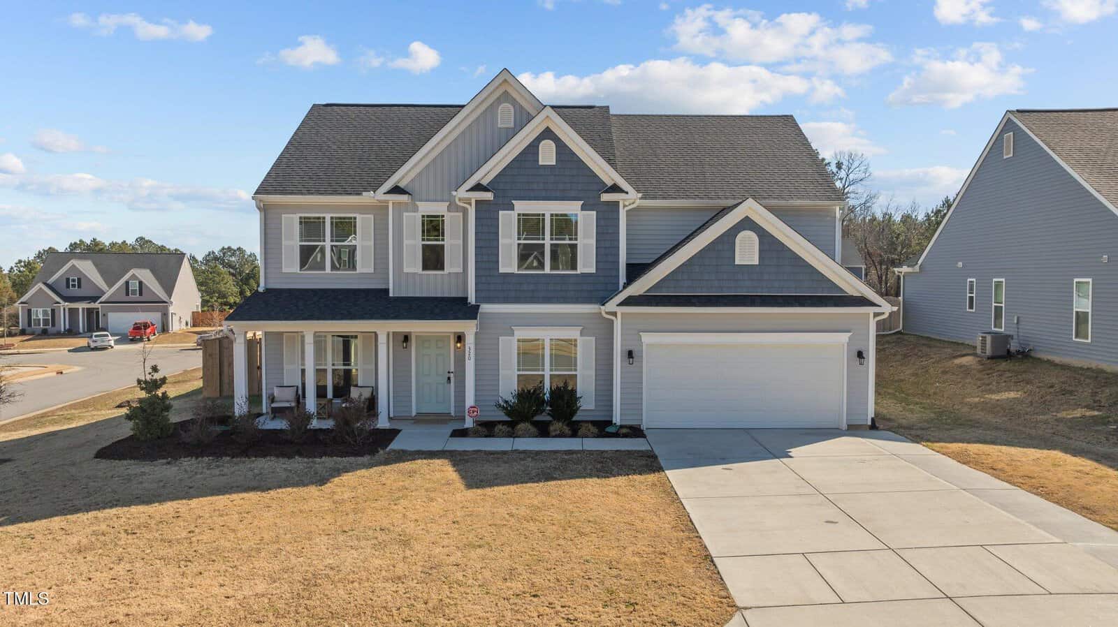 A two-story suburban house with gray siding, white trim, and a covered front porch. There is a double garage, a concrete driveway, and a lawn with minimal landscaping on a sunny day.