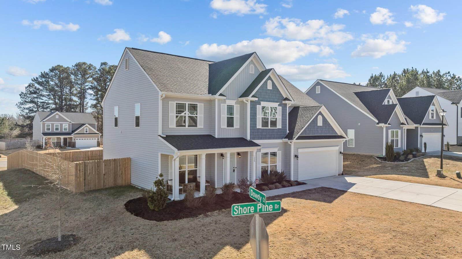 Two-story suburban house with gray siding and white trim, attached garage, small front porch, some landscaping, fenced backyard, and a street sign reading “Shore Pine Ln” at the corner. Neighboring houses visible in the background.