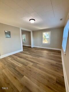 A vacant room with light-colored walls and hardwood flooring. The room features two windows on adjacent walls, a ceiling light fixture, and a small inset wall niche. Bright natural light illuminates the space.
