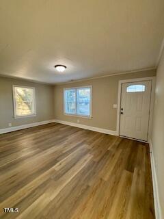 An empty living room with light wood flooring, beige walls, and a white ceiling. Two windows are on the far wall, and a white door is on the right. A ceiling light fixture is present.