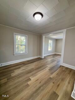 An empty room with light-colored walls and wood flooring. Two windows allow natural light in. A round ceiling light is centered above. An open entrance leads to an adjoining room with similar decor.