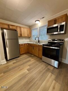 A compact kitchen with wooden cabinets and stainless steel appliances, including a refrigerator, microwave, and oven. The floor is wood and there is a double sink by two windows. White walls and a ceiling light fixture brighten the space.