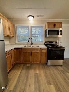 A kitchen with wooden cabinets, a stainless steel refrigerator, and an oven with a microwave above. There is a double sink under two windows, and a light fixture on the ceiling. The floor is wood-style laminate.