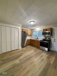 A kitchen with wooden cabinets, a stainless steel refrigerator, an oven with a microwave above, and a sink under two windows. The floor has light wood-style flooring, and there are louvered closet doors to the left.