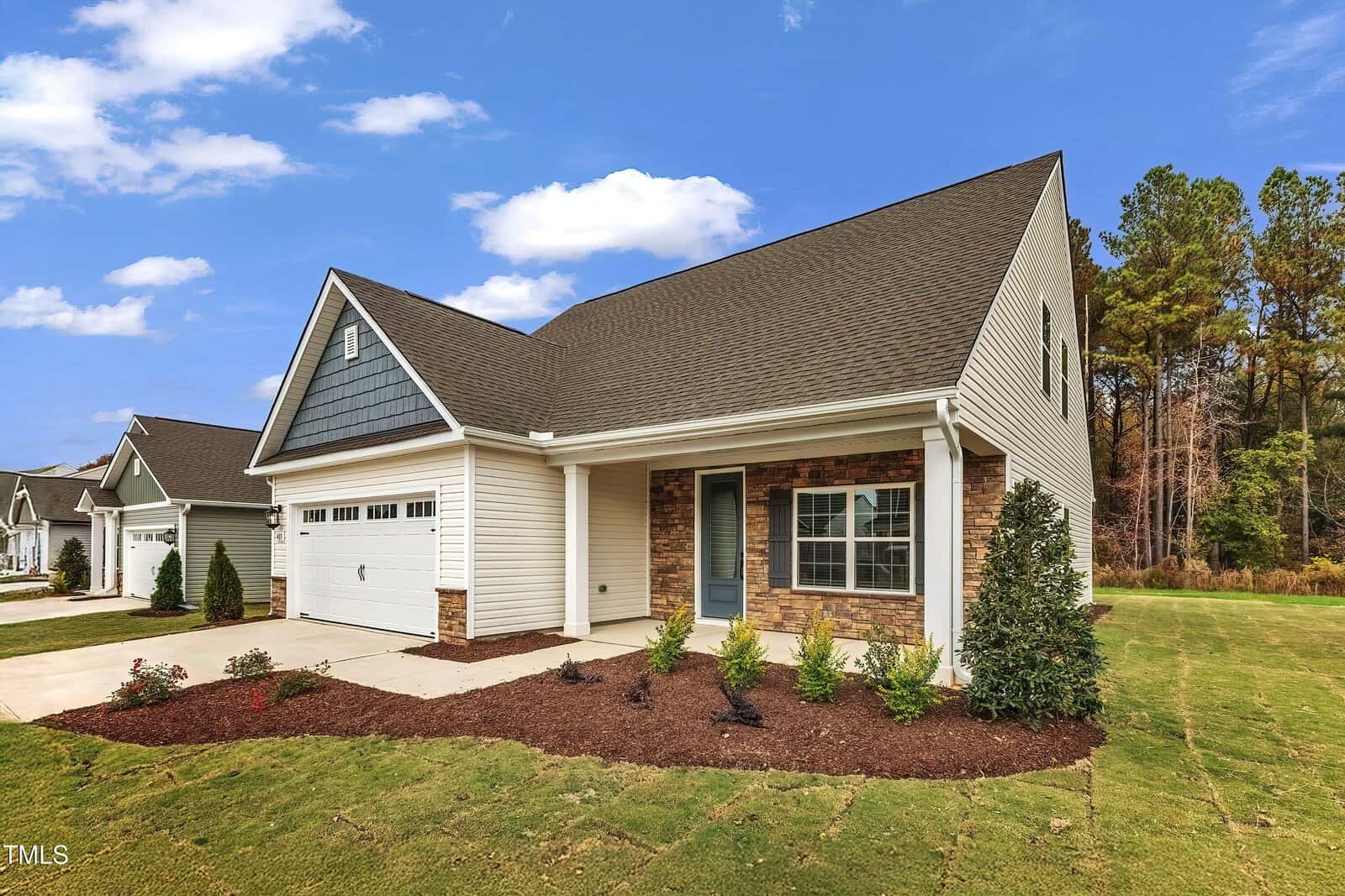 A single-story suburban house with a two-car garage, light-colored siding, dark roof shingles, and brick accents. There is a small front porch and neatly landscaped lawn with shrubs and mulch beds.